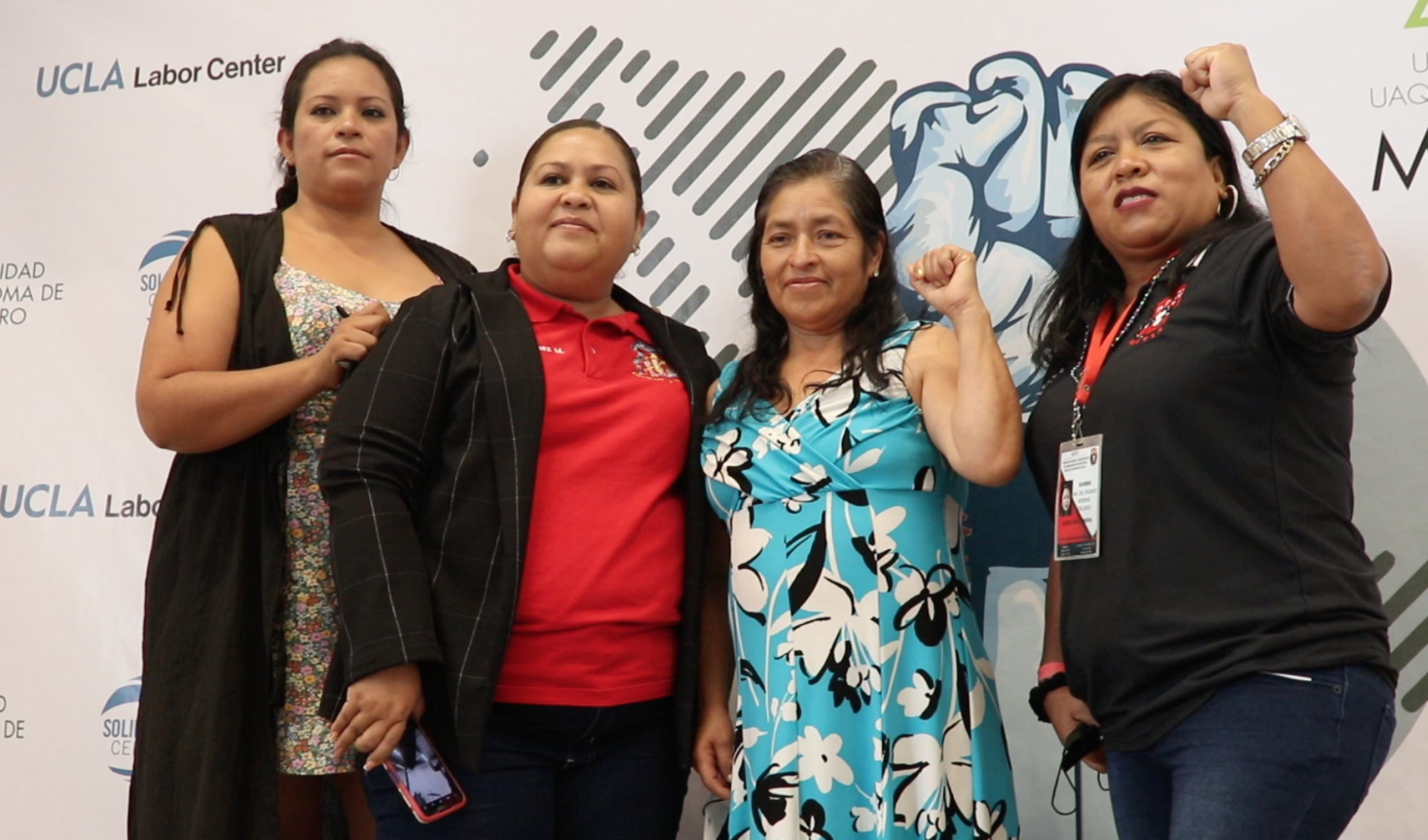 Four women stand and smile in front of a backdrop, two with raised fists.